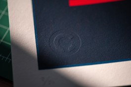 Simplicity debossed seal. Close-up of the debossed “Ç” logotype by Marçal Prats, impressed into the bottom-left corner of the navy blue print area of