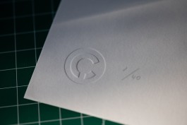 Marçal Prats' embossed monogram. A close-up crop of the lower left corner of a limited-edition print resting on a green grid-cutting mat. The photograph highlights a crisp blind emboss of a circular logo—two nested arcs with a small gap—pressed into thick white paper. Just to the right of the seal, in delicate pencil, is the artist’s edition notation “1/40.” The soft side lighting reveals the paper’s subtle texture and the raised relief of the emboss, suggesting the tactile care behind this hand-finished artwork.