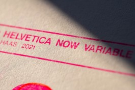 Typography (Technology) macro. Macro detail of the risograph print Typography (Technology) showing the text “Helvetica Now Variable – Haas 2021” in fluorescent pink ink. The close-up captures the subtle texture of the SeaWhite of Brighton paper and the slightly uneven ink coverage typical of risograph printing. A soft shadow cuts across the frame, enhancing the surface grain and revealing faint misregistration around the letter edges, adding to the analogue character of the piece.
