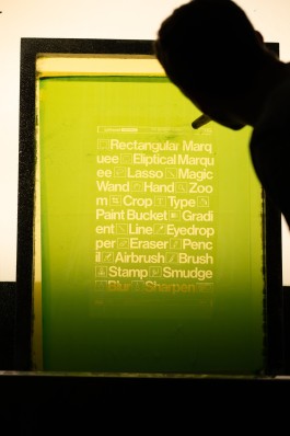 Design Tools screen preparation. Photograph of a silkscreen being washed out after exposure, revealing the design for an artwork based on classic graphic design tool icons and typography. A backlit green emulsion screen shows bold white text and pictograms from tools such as “Lasso”, “Crop”, “Brush”, “Smudge”, and “Sharpen”, arranged in a clean vertical layout. The silhouette of a person in the foreground directs a hose onto the screen. The composition is sharply defined, with warm light behind the translucent stencil highlighting the detail in the emulsion.