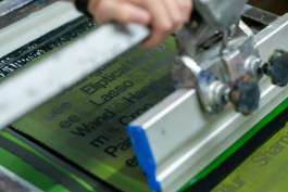 Production Tools process. Close-up of a screenprinting process in action. A squeegee glides across a green emulsion screen, pushing black ink through exposed areas that spell out words like “Lasso”, “Wand”, “Crop”, and “Sharpen” in bold sans-serif lettering. The partially printed design is visible underneath the mesh. The operator’s hand is slightly blurred in motion, capturing the dynamic act of inking. The detail highlights the mechanical precision of screenprinting and the clarity of the exposed stencil.