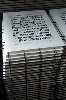 Design Tools stacked sheets. A freshly screenprinted poster rests on a drying rack. The print features a tightly composed list of digital graphic design tool names—such as “Lasso”, “Crop”, “Brush”, and “Smudge”—each paired with its corresponding icon. The type is bold and black, printed on smooth white paper, with sharp alignment and crisp edges. The layout mimics the interface of early design software, with text arranged in stacked lines and neat groupings. Surrounding prints are visible underneath in the rack of theproduction run in progress.