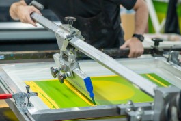 Interdisciplinarity process 02. Close-up of a screenprinting process in action, showing a bright yellow-green ink layer being pulled across a silkscreen using a mechanical press. This is the third colour pass of