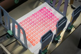 Typography (Technology) risograph. This image captures a freshly printed risograph sheet of Typography (Technology) exiting a risograph printer. The design features a repeated grid of lowercase “a” characters transitioning in weight and size from top to bottom. The fluorescent pink and orange inks produce a vivid gradient effect across the page. The risograph’s feed tray and guide arms are visible, framing the print as it finishes. The detail highlights both the mechanical printing process and the texture of the risograph ink on paper.