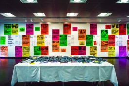 CCD KSA Exhibit General View. A vibrant exhibition space featuring the River Rights: Guardians for the Hogsmill project. A large wall is covered with neon posters in pink, orange, green, yellow, and purple, displaying urgent headlines on UK river pollution and environmental justice. In front, a long table with a white cloth displays folded publications and a central textile artwork made from indigo-dyed patches, reflecting themes of restoration and community care.