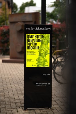 A bright yellow event poster displayed outdoors at the Stanley Picker Gallery. The poster, titled “River Rights: Guardians for the Hogsmill,” features bold typography and an abstract woven pattern, placed on a sleek, black sign. The background shows blurred bicycles and greenery, giving context to an urban gallery setting.