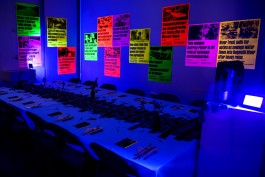 SPG River Rights Dinner Room. A dimly lit room with a long, formally set dinner table under blue lighting, surrounded by neon posters on the walls highlighting UK river pollution headlines. The posters are in fluorescent colours—pink, yellow, green, and orange—and feature bold text from environmental news reports. The event setting evokes urgency around river rights and environmental justice, combining activism with communal gathering.