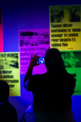 SPG River Rights Event. A visitor is silhouetted in blue and purple lighting while photographing a wall of fluorescent posters highlighting UK river pollution issues. The posters, in neon yellow, green, and magenta, display bold headlines about sewage spills and water contamination. The scene captures public engagement with environmental activism in a visually striking, immersive installation.