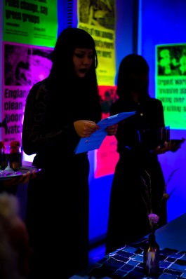SPG River Rights Event. SPG River Rights Event. A group of people sit around a long table in a dark room illuminated by deep blue light. The walls are covered with fluorescent posters in neon orange, pink, yellow, and green, displaying alarming headlines about UK river pollution and environmental degradation. A small screen shows footage of a river, reinforcing the theme. The scene combines visual activism and participatory engagement in an immersive, awareness-raising installation.