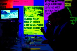 SPG River Rights Drinks. Guests seated at a long table sip from glasses under glowing blue and violet light. A nearby screen shows river footage, while fluorescent posters dominate the background, with headlines like “Thames Water fined £4 million after catastrophic sewage blunder.” The scene is part of an immersive dinner installation confronting water pollution through sensory experience and environmental storytelling.