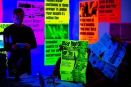 SPG River Rights Booklet Present. An attendee stands and reads from a leaflet under deep blue lighting, while others examine printed materials at a dinner event focused on river advocacy. One guest holds a large neon green poster from the booklet reading “River Rights: Guardians for the Hogsmill.” Behind them, fluorescent posters highlight environmental headlines about UK river pollution. The immersive setting blends activism, information, and communal dialogue.