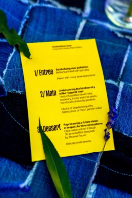 SPG River Rights Menu. A close-up of a vibrant yellow curated dinner menu placed on indigo-dyed patchwork fabric. The menu outlines a three-course meal inspired by themes of river pollution and ecological restoration: a spirulina-tinted nettle tea for the entrée, a herb-infused main highlighting local biodiversity, and a dessert symbolising future water purity, served with filtered water and chalk sweets. Dried leaves and lavender add a natural, thematic touch.