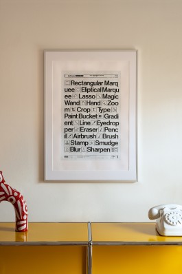 Design Tools framed. Framed on a neutral-toned wall, the artwork Design Tools is presented with clean, contemporary styling. The piece is centred in a white mount and a minimal white frame with a slim profile, creating a stark contrast that highlights the crispness of the black text and graphics. Below, a yellow gloss cabinet and vintage white rotary telephone introduce nostalgic accents that complement the artwork’s references to analogue design tools. The photograph is evenly lit, allowing the hand-applied varnish finish to subtly catch natural light, giving depth to the printed surface.