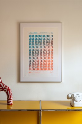 Typography (Technology) Blue framed. This image displays the “Typography (Technology)” artwork by Marçal Prats, framed and hung on a beige wall above a contemporary yellow storage unit. The print itself depicts repeated lowercase “a” characters arranged in rows, transitioning from aqua blue at the top to vibrant orange at the bottom, closely resembling traditional Letraset rub-on transfer sheets. The minimalist white frame and clean mounting complement the modern decor, highlighted by a sculptural red-and-white ceramic piece to the left and a retro-style white telephone to the right, creating a balanced visual arrangement that accentuates the blend of nostalgia and contemporary graphic exploration in the artwork.